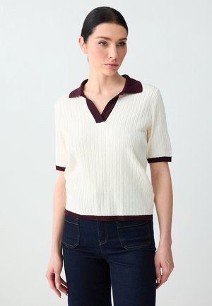 Donna con polo di maglia bianca a maniche corte con colletto e polsini scuri, abbinata a jeans blu scuro, in piedi davanti a uno sfondo semplice.