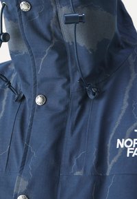 Tmavě modrá nepromokavá bunda s texturovaným povrchem, nastavitelnými šňůrkami, zapínáním na kovové knoflíky a logem na přední straně.