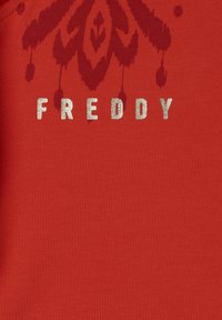 Camiseta de tela roja con un patrón floral texturizado y el texto "FREDDY" en letras metálicas plateadas. Diseño clásico de cuello redondo.