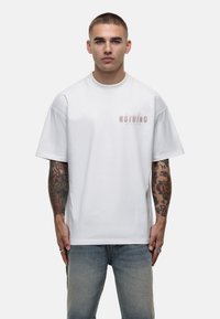 Witte katoenen t-shirt met korte mouwen en een ronde halslijn. Voorzien van een lichtroze bedrukte tekst "NOTHING" aan de voorkant. Standaard pasvorm.