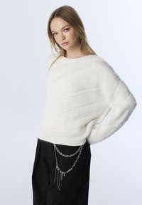 Pull en maille blanche texturée avec des rayures diagonales, décolleté large et poignets côtelés. Associé avec un pantalon noir avec des accents en chaîne argentée.