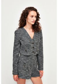 Chaqueta corta de tweed gris con diseño de doble botonadura, botones en tono dorado, y pantalones cortos a juego con bolsillos delanteros y tejido texturizado.