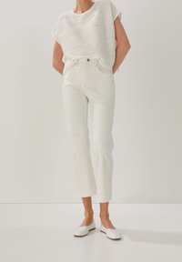 Kvinde iført struktureret hvid kortærmet bluse, højtaljede lige hvide jeans og hvide flade sko mod en ensfarvet baggrund.