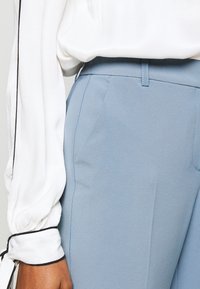 Närbild av ljusblå skräddarsydda byxor med en slät textur, med sidofickor. En vit blus med rynkade ärmar och svarta detaljer syns.