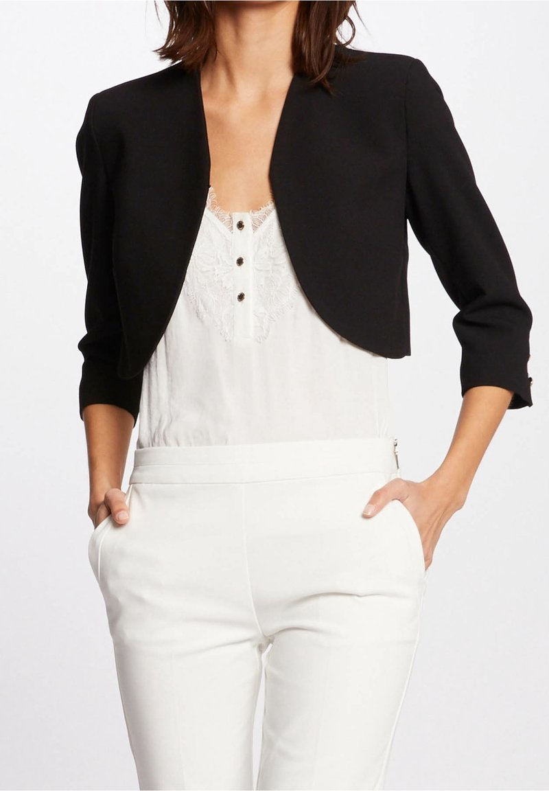 Woman wearing a black cropped blazer over a white lace-trimmed blouse and high-waisted white pants, hands in pockets.