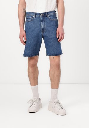 Jeansshorts i mellanmörk blå, med rak skärning, rå kant, sidfickor och knappstängning fram, kombinerat med vita sneakers och strumpor.