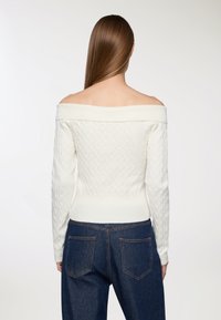 Sweter w kolorze off-white, dziergany, z designerskimi ramionami, żeberkowym dekoltem i dopasowaną talią. Noszony z ciemnoniebieskimi spodniami o wysokim stanie.