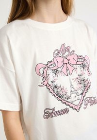 Vit bomullströja med en rosa hj�ärt-design, en rosett och en illustration av Eiffeltornet omgiven av rosa blommiga element.