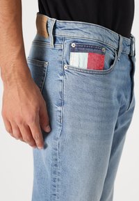 Blugi din denim bleu deschis, cu o croială slim, având o suprafață texturată și un lucru de patchwork în buzunarul din spate, cu accente în roșu, albastru și verde mentă.
