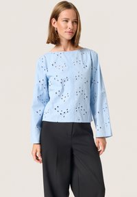 Ljusblå kort blus med texturerad yta och hålmönster, med långa �ärmar och vid halsringning. Tillsammans med svarta byxor.