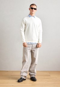 White cotton sweatshirt over a light blue collared shirt, gray denim pants, and black boots. Sunglasses complete the outfit.