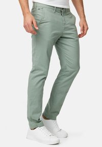 Pantalones de algodón verde claro con corte slim, trabillas para cinturón, bolsillos frontales y cierre de botón, combinados con zapatillas blancas.
