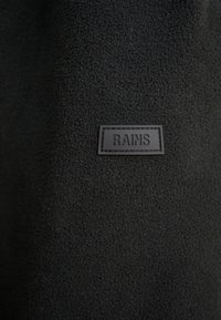 Tissu polaire noir avec une surface texturée et une pièce rectangulaire en cuir noir cousue, étiquetée "RAINS".