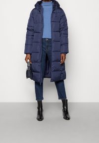 Marinblå puffjacka med huva, med kviltade sektioner, sidofickor och helfront. Kombinerad med en ljusblå kabelstickad tröja och jeans.