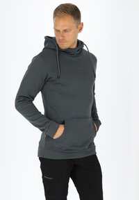 Felpa grigia in pile con tasca frontale, cordini regolabili e un piccolo logo sul petto. Abbinata a pantaloni neri per un look casual.