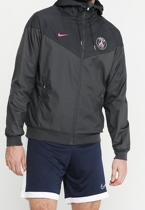 Man draagt een zwarte en donkergrijze sportjas met rits, met het logo van Paris Saint-Germain en een roze Nike-swoosh, gecombineerd met marineblauwe Nike-shorts.