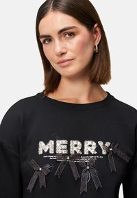 Czarna bluza z podniesionym napisem "MERRY" wykonanym z kryształków, z ciemnymi wstążkami i ozdobami z przodu. Okrągły dekolt, luźny krój.
