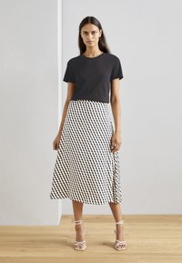 Zwart T-shirt met korte mouwen gecombineerd met een lange, witte rok met een geometrisch zwart patroon. Het model draagt strappy witte hakken.