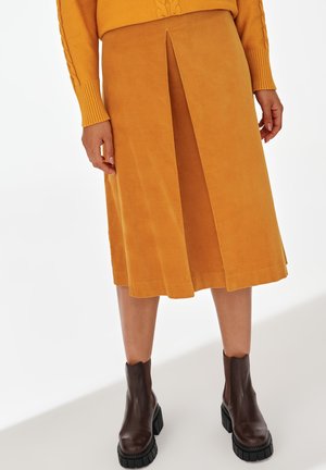Musztardowożółta spódnica midi z plisą z przodu, zestawiona z pasującym musztardowym swetrem i ciemnobrązowymi masywnymi botkami za kostkę.
