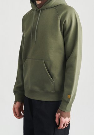 Mann trägt olivgrünen Kapuzenpullover mit Fronttasche und Kordeln, kombiniert mit schwarzer Hose vor neutralem Hintergrund.