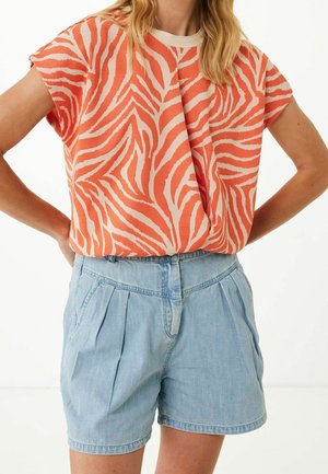 Femme portant un haut à manches courtes orange et beige à motif zèbre, rentré dans un short en denim bleu clair taille haute avec des plis.