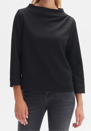 Kobieta ubrana w czarną, fakturowaną bluzkę z długim rękawem i dekoltem typu łódka oraz dopasowane ciemnoszare dżinsy, widoczna od brody do bioder.