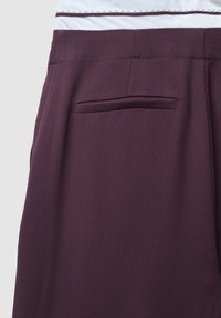Bordeauxfarbene gewebte Hose mit glatter Textur, ausgestattet mit einer Gesäßtasche und einem taillierten Schnitt. Dezente tonale Nähte an der Taille.