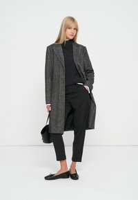 Cappotto herringbone grigio con rever, indossato sopra un dolcevita nero e pantaloni scuri, accessoriato con una borsa nera e ballerine.