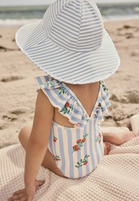 Blau-weiß gestreifter Sonnenhut mit fransigem Rand, getragen mit einem floralem Einteiler mit Rüschenträgern, sitzt am Sandstrand.