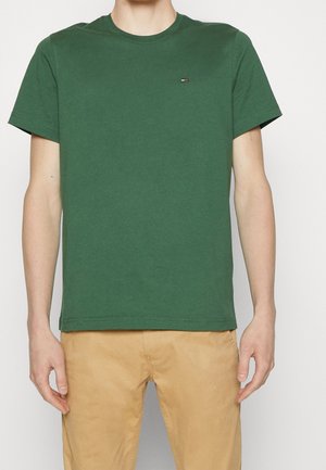 Homme portant un t-shirt vert uni à manches courtes et col rond et un pantalon beige, debout devant un fond clair.