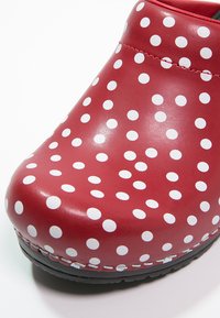 Zoccolo rosso con pois bianchi, realizzato in pelle lucida. Presenta la punta arrotondata e una suola nera con dettagli di cucitura visibili.