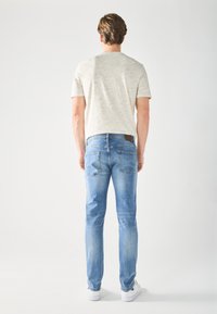 Homme debout de dos, portant un t-shirt beige clair, un jean bleu et des baskets blanches sur un fond blanc uni.