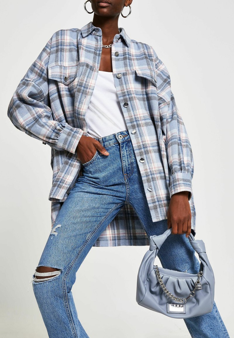 Femme portant une chemise à carreaux bleue surdimensionnée, un débardeur blanc, un jean bleu déchiré, tenant un sac à main bleu clair avec détail chaîne.