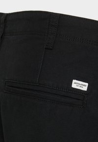 Crne traperice od denima s stražnjim džepom s skrivenim patentnim zatvaračem. Bijela oznaka s natpisom "JACK & JONES EST. 1990" je prišivena.