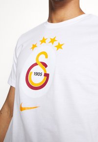 Camicia bianca con logo Galatasaray giallo e rosso che presenta cinque stelle gialle e "1905", con uno swoosh arancione Nike sottostante.