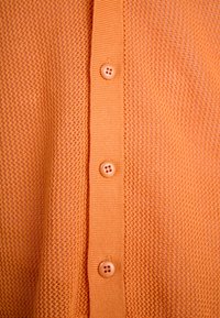 Helles orange Strickcardigan mit einem strukturierten Muster, sechs Knöpfen auf der Vorderseite und gerippten Akzenten entlang der Knopfleiste.