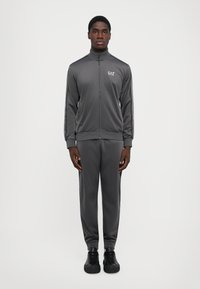 Chándal de hombre en gris oscuro, con una chaqueta con cremallera y cuello alto, adornada con rayas laterales, combinada con pantalones ajustados a juego y zapatillas negras.