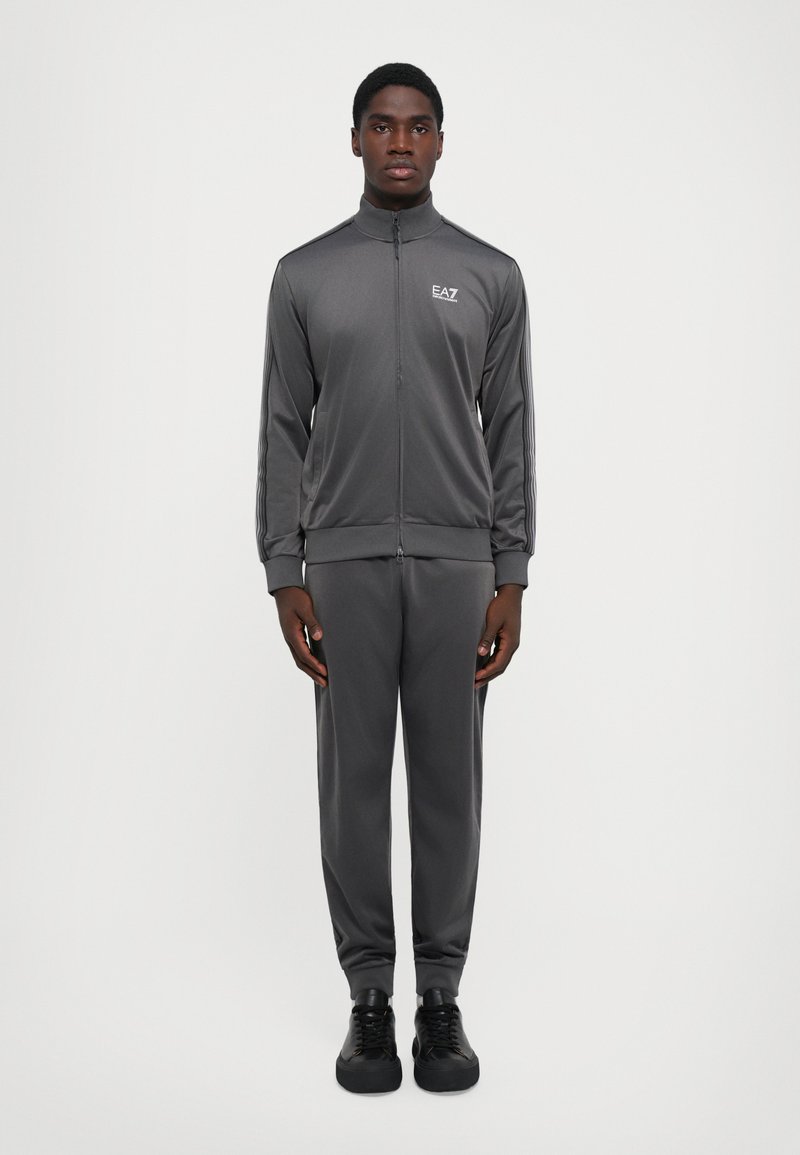 Chándal de hombre en gris oscuro, con una chaqueta con cremallera y cuello alto, adornada con rayas laterales, combinada con pantalones ajustados a juego y zapatillas negras.