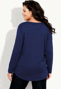 Granatowa bluzka z długim rękawem, zaokrąglonym dołem i okrągłym dekoltem. Miękki, fakturowany materiał. Modelka pokazana od tyłu, w ciemnych jeansach.