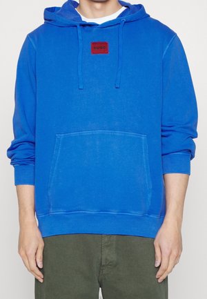 Blauwe katoenen hoodie met een voorzak, ribgebreide mouwen en een vierkante rode logopatch met de tekst "HUGO" op de borst. Glad textuur.