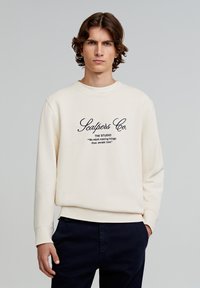 Beige sweatshirt met zwarte tekst in cursief. Heeft een ronde halslijn, geribde manchetten en zoom. Draagt, met donkere broek, handen in de zakken.