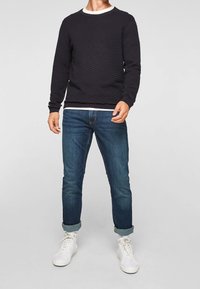 Granatowy sweter o fakturze z prążkowanym dekoltem, zestawiony z niebieskimi dżinsami i białymi trampkami. Sweter ma poziome żłobienia dla dodatkowego efektu.