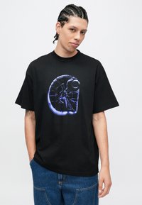 Svart bomulls-T-skjorte med en stor, belyst grafisk logo på forsiden. Korte ermer, avslappet passform, med rund hals.