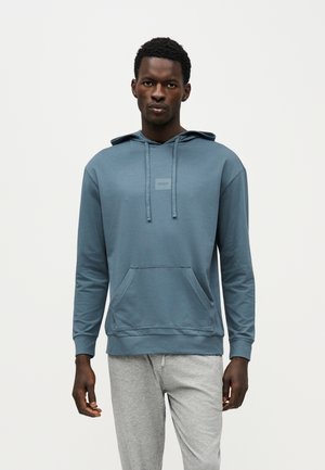 Un homme porte un sweat à capuche bleu avec une poche avant et un pantalon de jogging gris, sur un fond clair et uni.