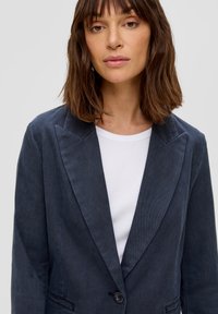 Granatowy żakiet z korduroju z szerokimi klapami, zapinany na pojedynczy guzik, z dwoma przednimi kieszeniami, noszony na białym t-shircie z okrągłym dekoltem.