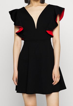 Femme portant une robe noire sans manches avec un décolleté en V profond et des épaules volantées doublées de rouge, debout devant un fond uni.