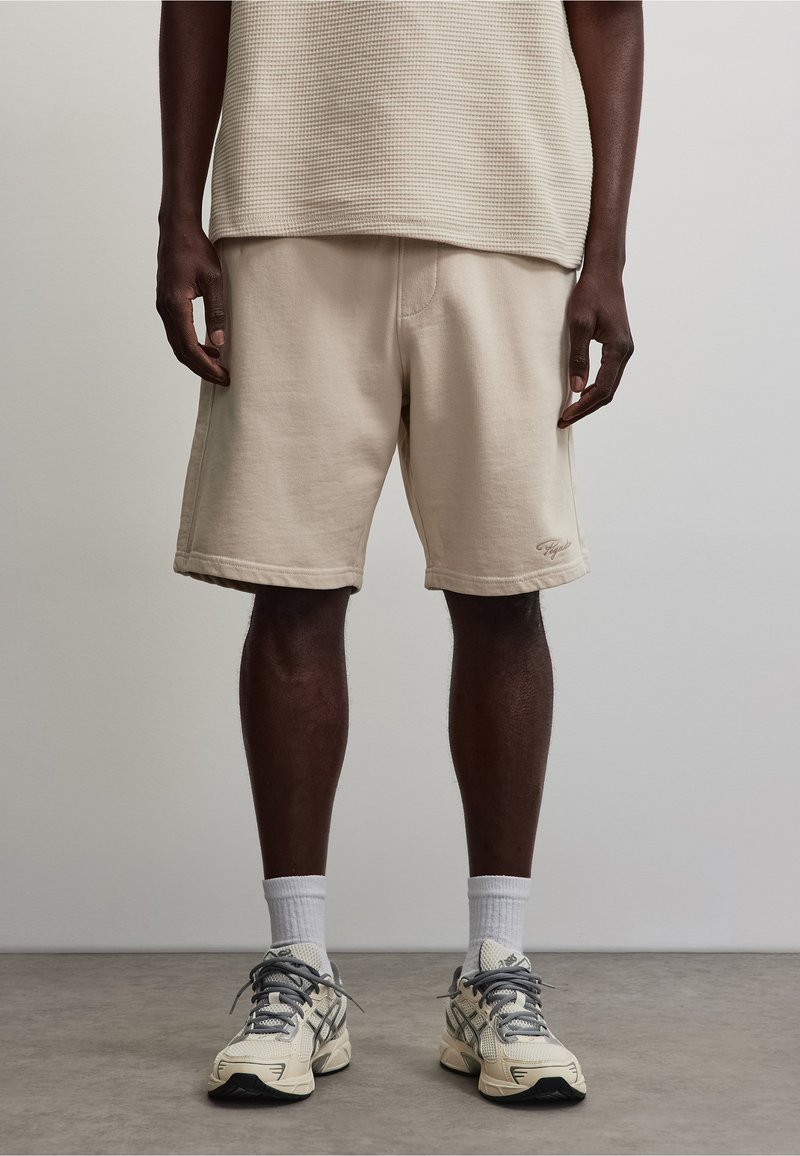 Homme portant un short beige, des chaussettes blanches et des chaussures de course crème, debout sur un sol gris devant un fond uni.