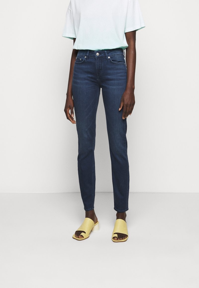 Donkerblauwe slim-fit jeans met een gladde textuur, gecombineerd met een lichtgroen t-shirt en gele sandaaltjes met open tenen op een neutrale achtergrond.