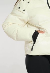 Crème-kleurige pufferjack met een glanzende afwerking, voorzien van zwarte accenten, drukknoopsluitingen en een zijzak met rits.