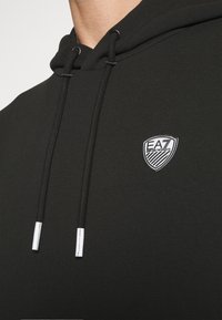 Svart hoodie i mjukt tyg, med en dragsko på huvan med silverfärgad spets och en liten EA7-logotyp på br östet.